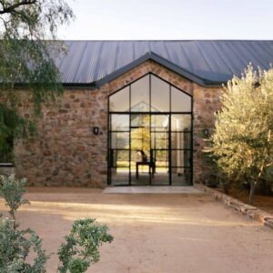 Entrada del lodge con muros de piedra, un alto hastial acristalado de marco negro, tejado de chapa ondulada, olivos y un patio de llegada de grava rastrillada