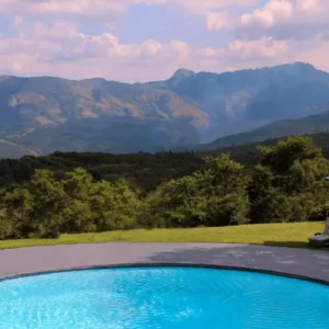 Piscina circular azul com pavimento à volta, espreguiçadeiras às riscas e dois guarda-sóis no relvado, com uma cordilheira florestada ao fundo