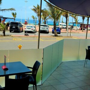 Mesas exteriores bajo un toldo en Clippers Café, frente a Marine Parade y la playa