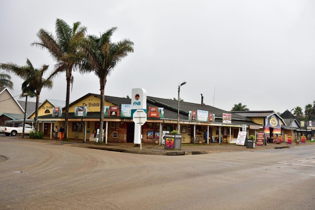 A row of low verandahed curio and craft shops behind tall palms on an empty tarred street corner, hand-painted signboards outside