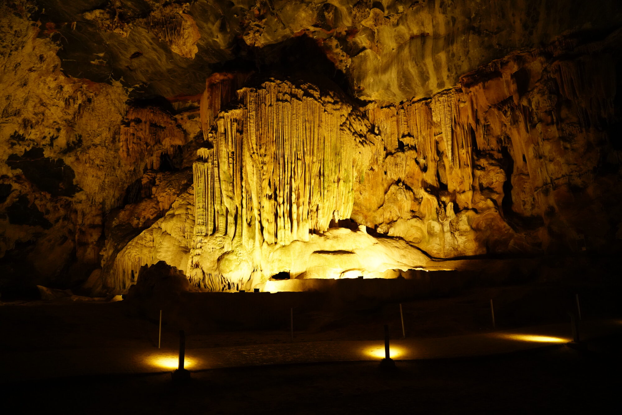 A vast underground chamber with amber-lit flowstone curtains and stalactite columns rising above a dark cave floor