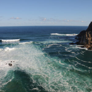 Surf and turquoise water surging through the channel between the Knysna Heads, with the rocky eastern headland cliff and open ocean beyond