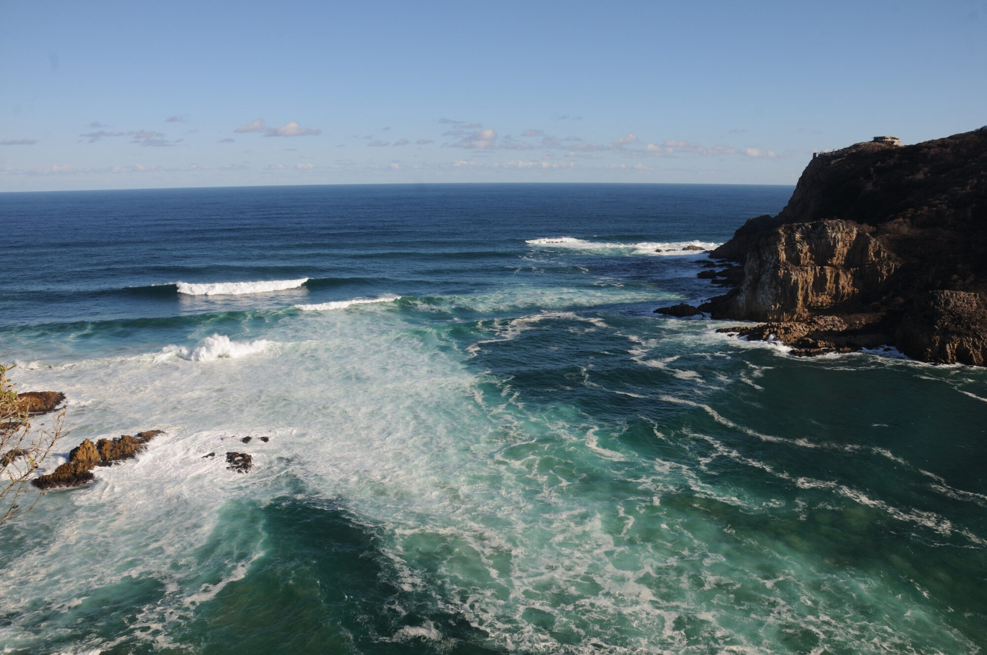 Surf and turquoise water surging through the channel between the Knysna Heads, with the rocky eastern headland cliff and open ocean beyond