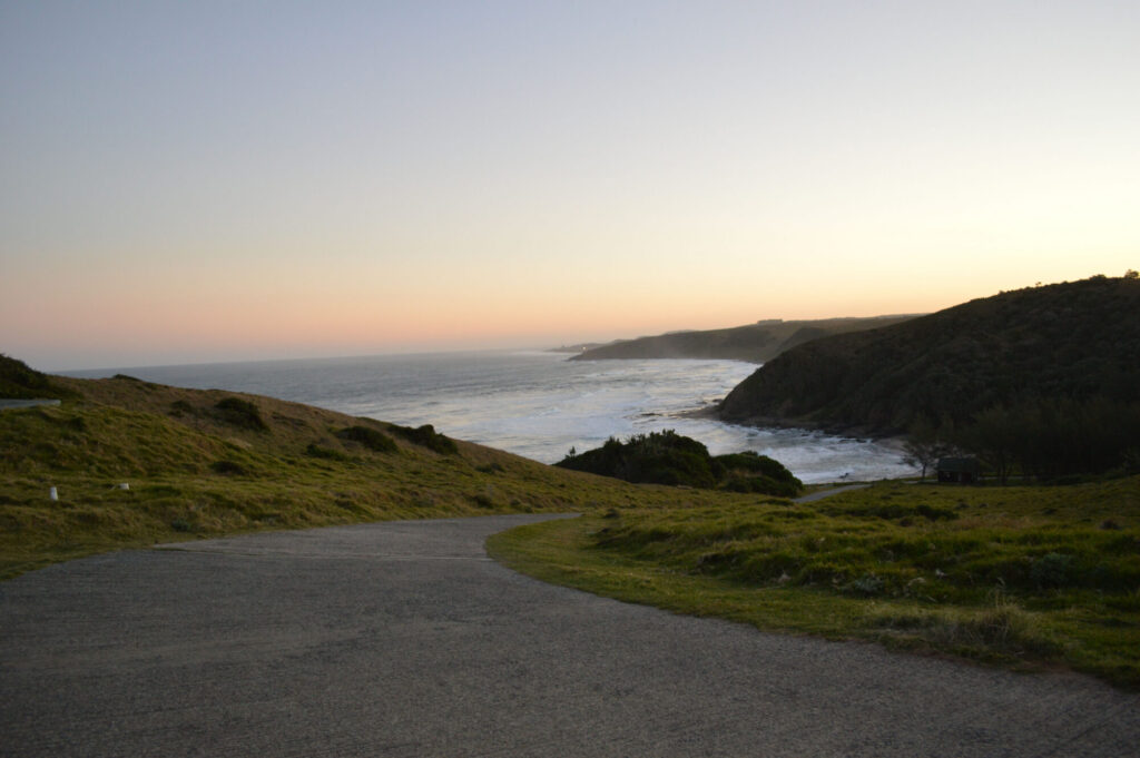 A tarred road curving down grassy coastal headlands towards a surf-lined bay and distant cliffs at sunset on the Eastern Cape coast