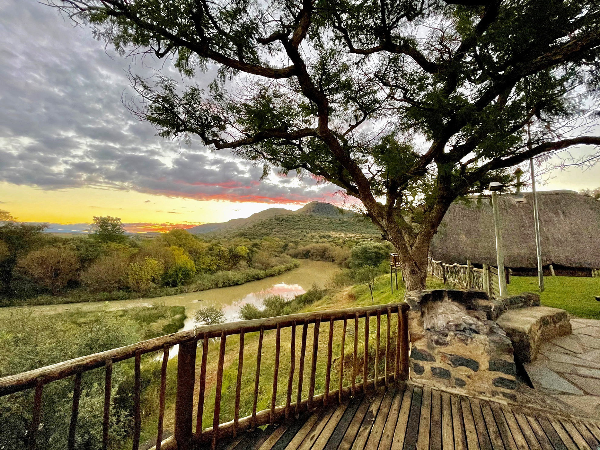 Vue depuis la terrasse en bois du lodge, au-delà d’un grand arbre, sur la Tugela River et les collines au coucher du soleil, avec le toit de chaume du lodge à droite