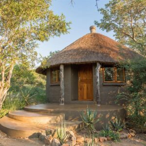 Round grey-plastered hut with a conical thatched roof, timber door and raised concrete step-up terrace, set among bush and aloes